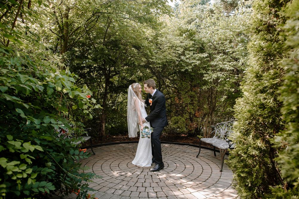 A couple shares their first moments as newlyweds on a circular stone patio surrounded by lush summer greenery.