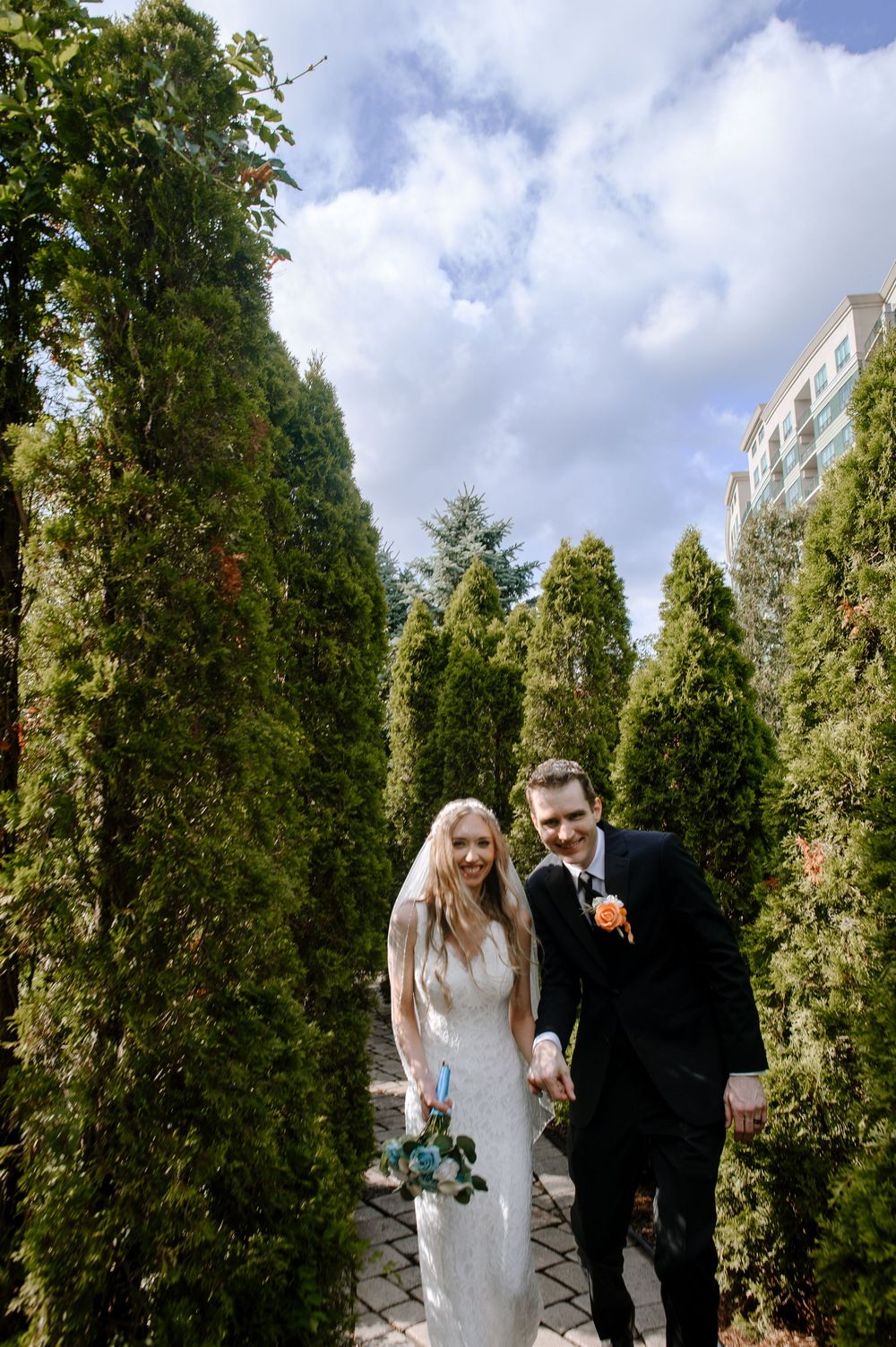 A wedding couple walks hand in hand along a garden path lined with tall evergreen trees.