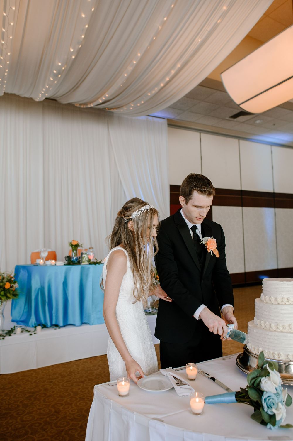 Wedding couple cutting their elegant white tiered cake together at a reception decorated with blue accents and white draping.
