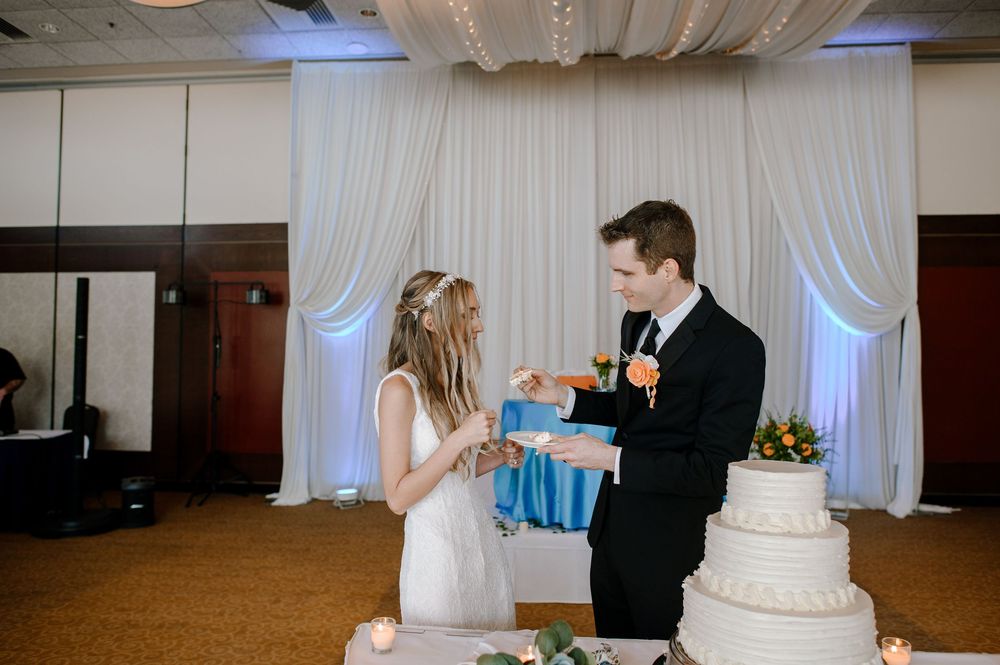 A couple in wedding attire share a playful cake feeding moment in front of white drapes and a tiered cake at their reception.