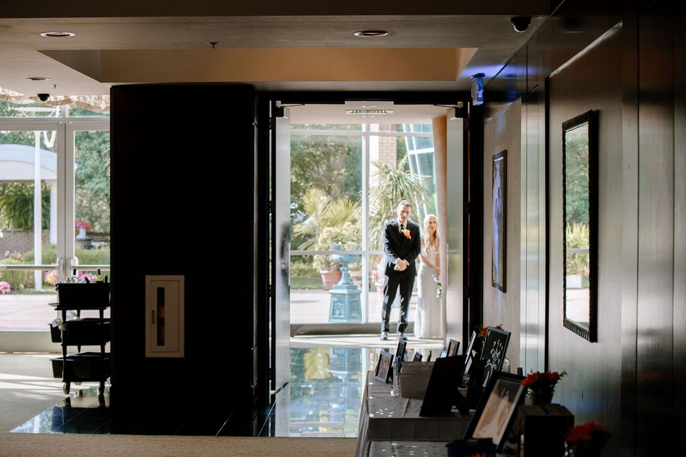 Modern restaurant interior with sunlit windows, dark walls, and wooden chairs arranged along a mirrored wall.
