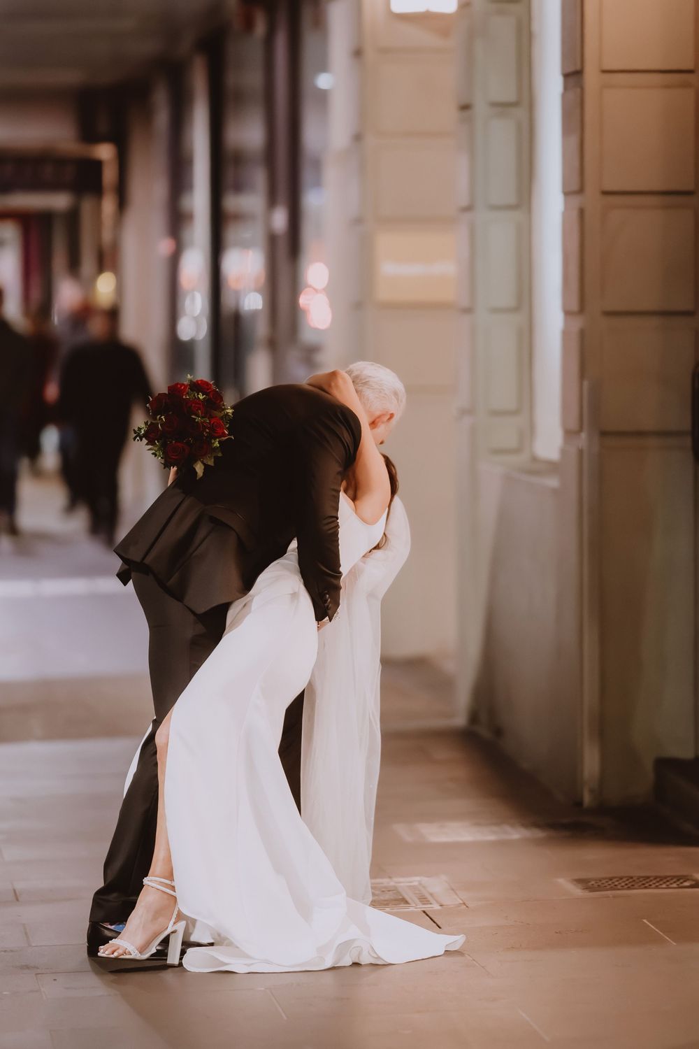 A couple in wedding attire dances romantically in an elegant hallway with stone columns and warm lighting.