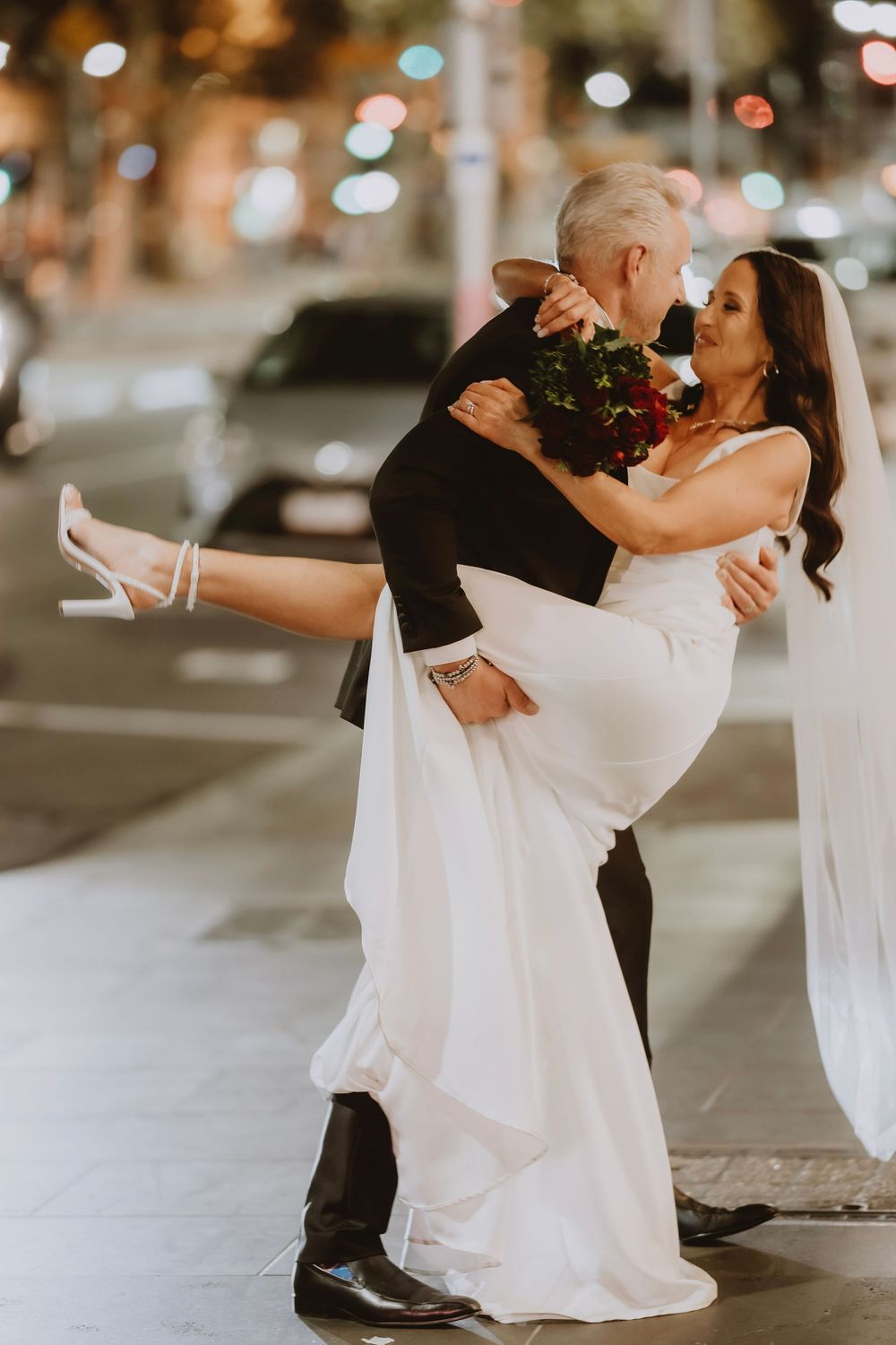 A couple in formal wedding attire dances intimately on a city street at night with blurred lights in the background.