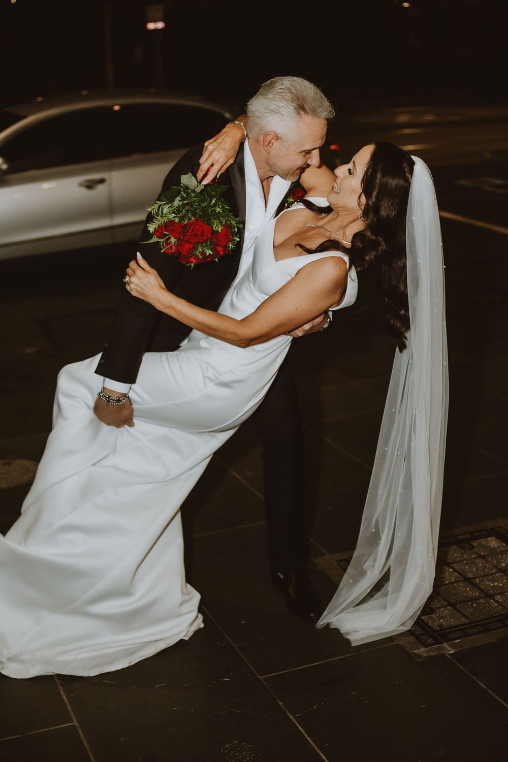 A bride in a flowing white wedding dress with veil gracefully dips and spins across a dark floor during a dance.