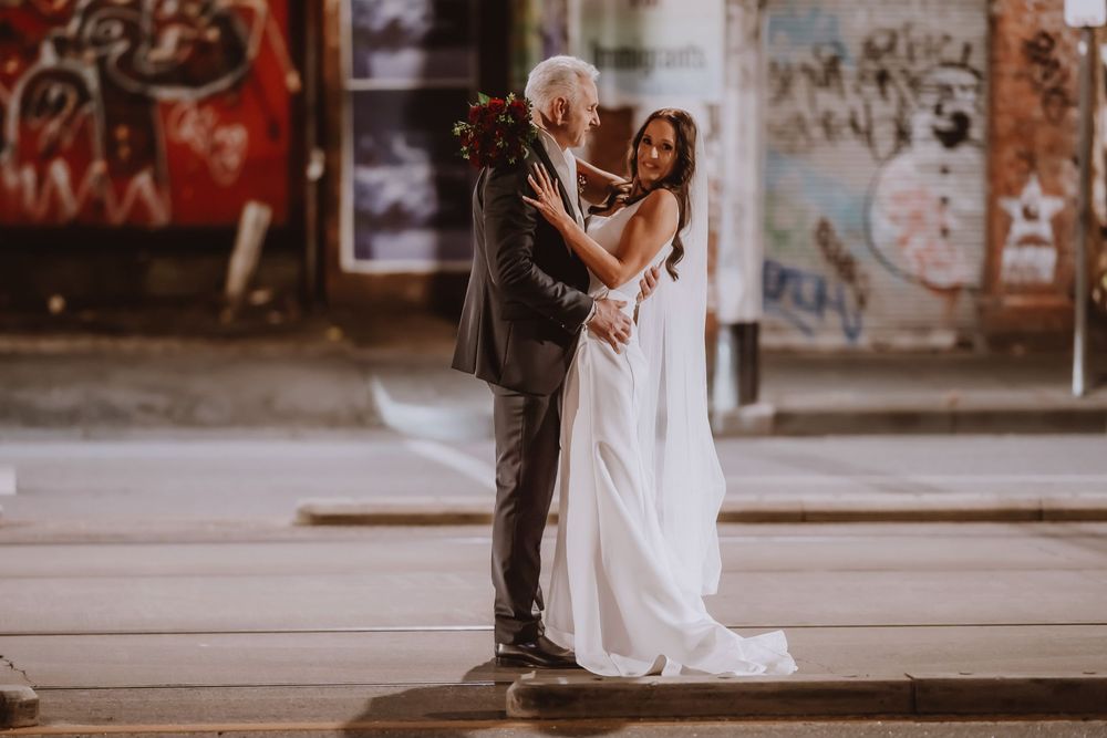 A couple in wedding attire shares a sweet embrace against an urban brick wall backdrop with graffiti.