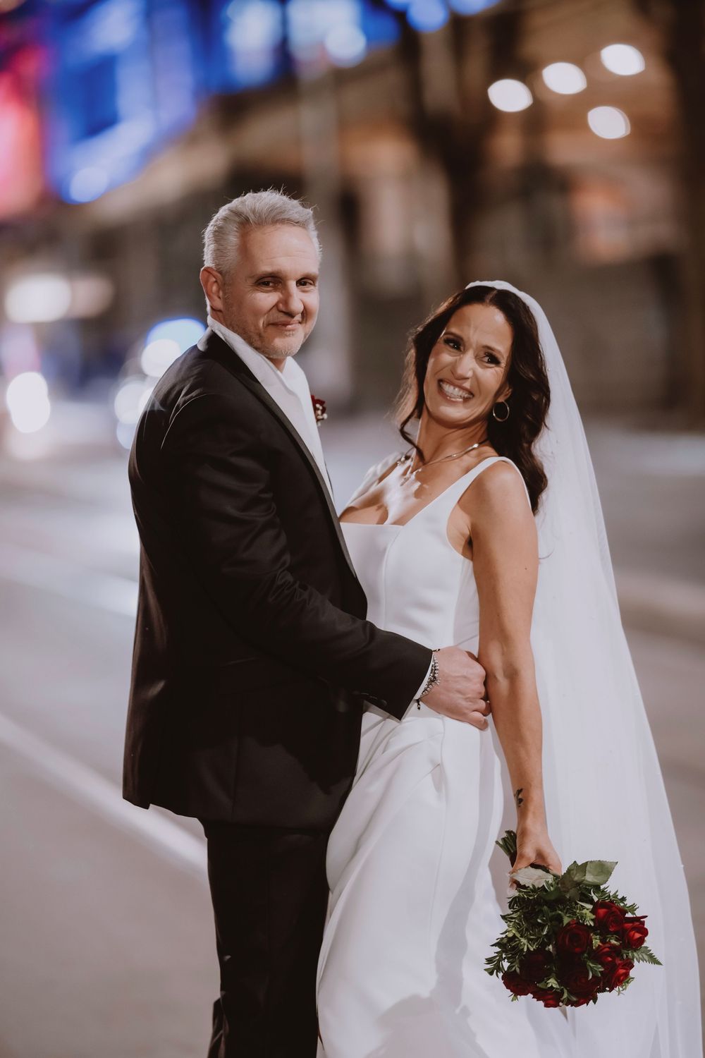 A bride and groom share intimate moments in an urban setting with a bouquet of deep red flowers.