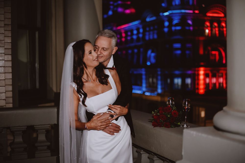 Romantic wedding couple embraces in front of colorful nighttime city lights through a window.