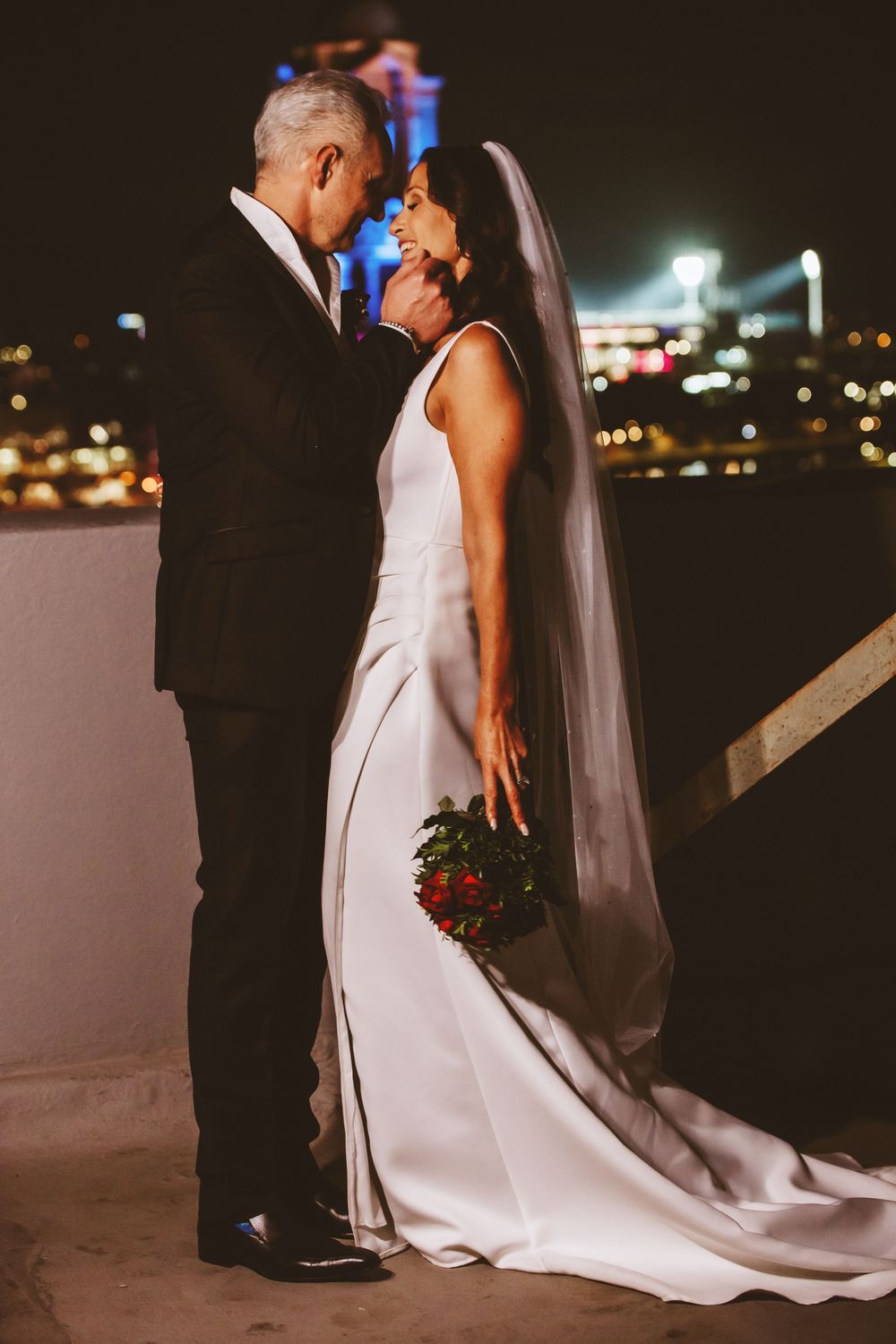 Romantic wedding couple embraces at night with city lights glowing in the background creating a dreamy atmosphere.