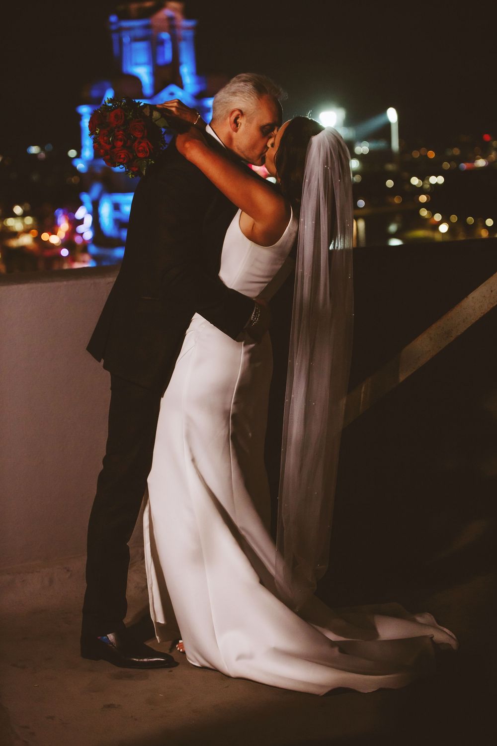 A bride in a white dress and groom in black dance together on a rooftop terrace at night with city lights glowing behind them.