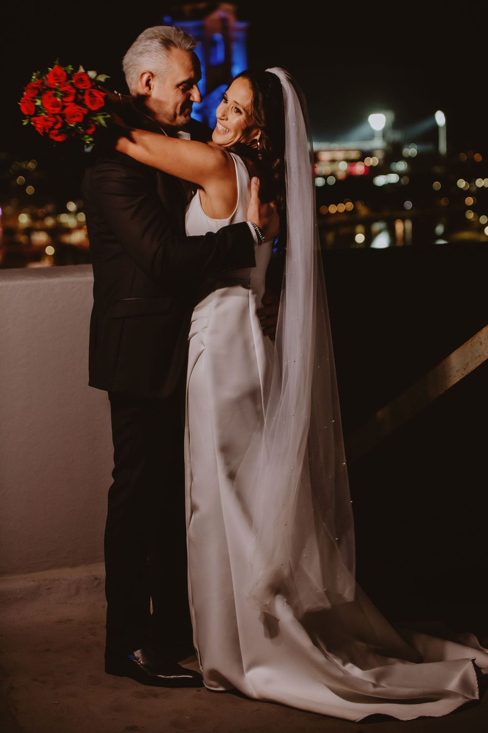 Elegant night wedding portrait with bride in flowing white gown and dramatic city backdrop.