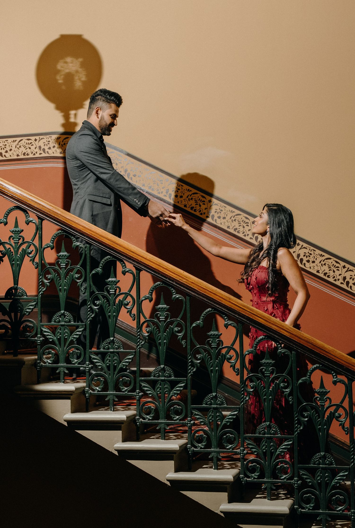 Couple in formal attire on an ornate staircase with elegant dark green railings creates romantic moment during photoshoot.