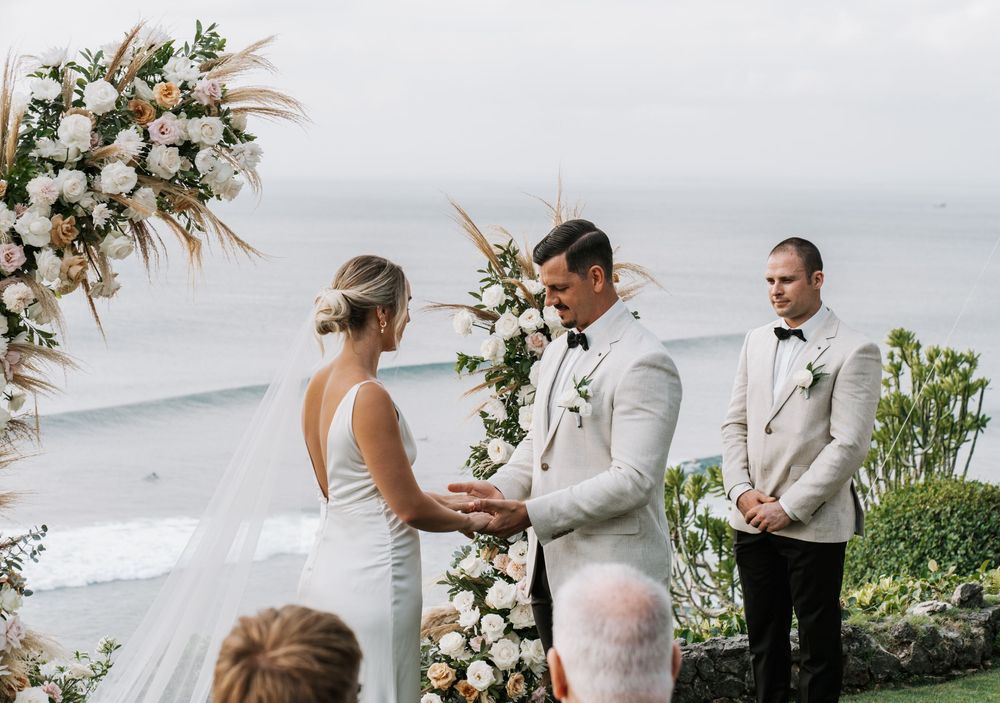 Romantic beach wedding ceremony taking place on a scenic coastline with tropical greenery and white floral arrangements.