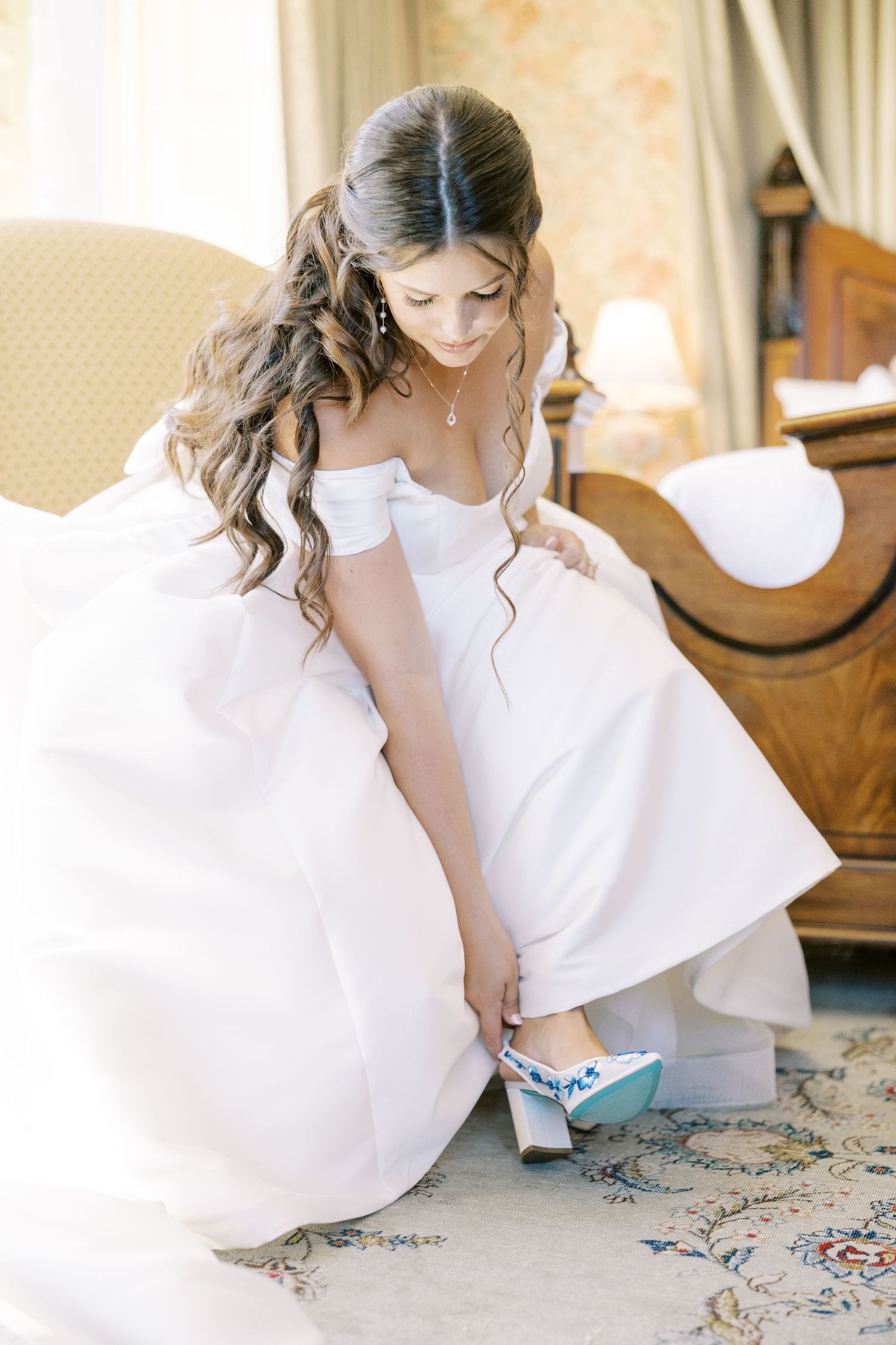 Someone in a flowing white dress sits gracefully on an ornate rug in a sunlit room.