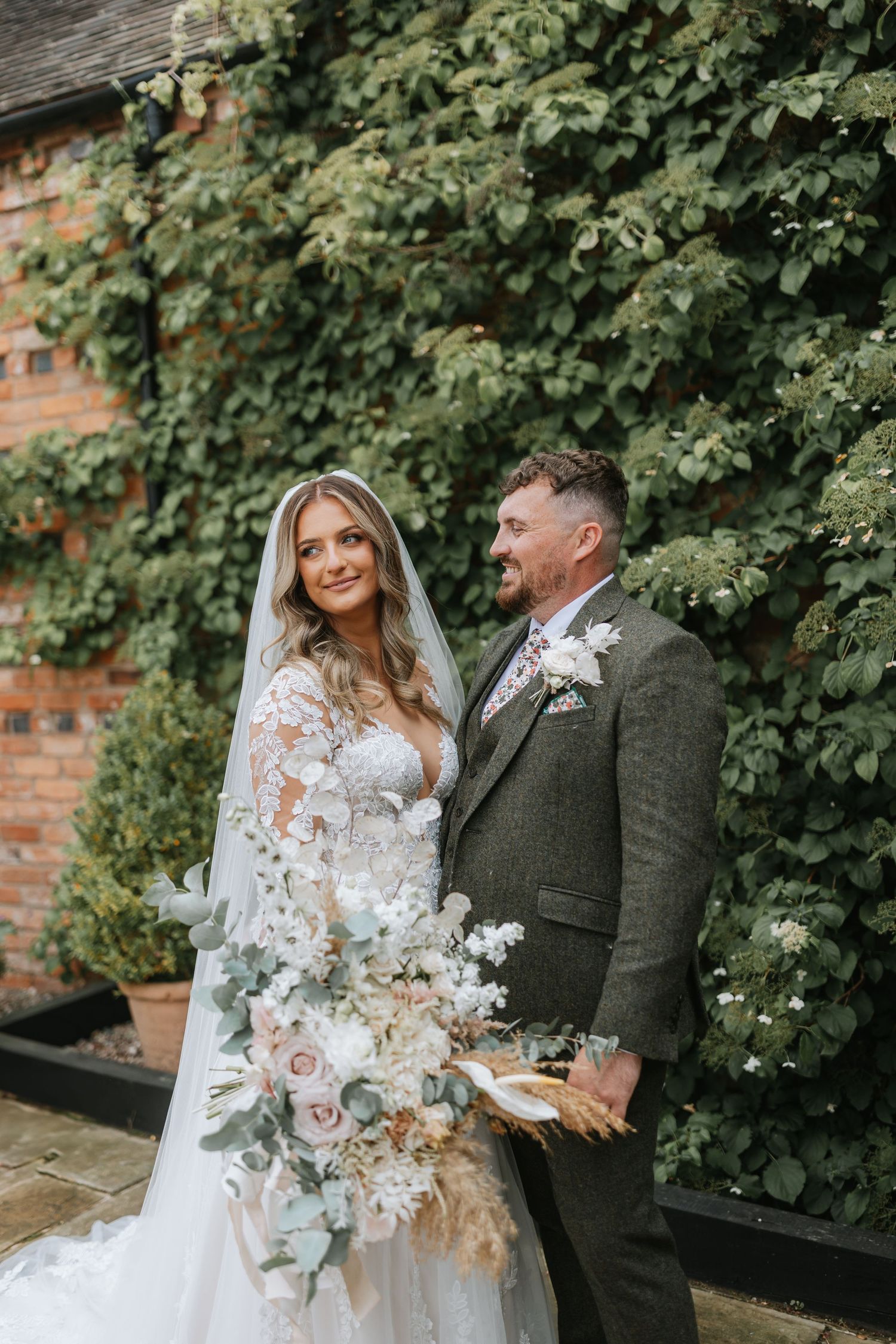 A couple shares joyful moments against an ivy-covered brick wall on their wedding day in formal attire.