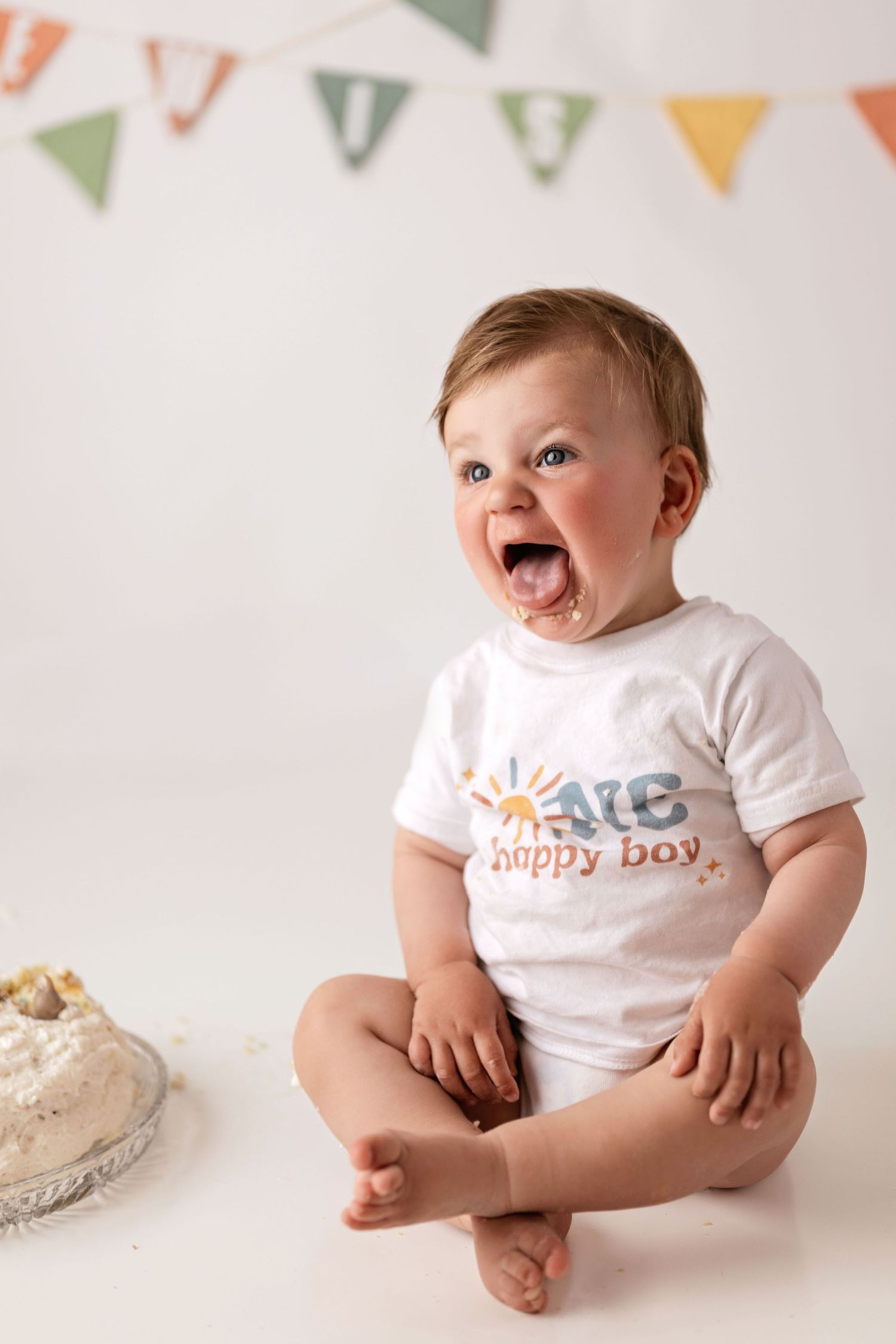 A baby wearing cute graphic t-shirts sits on white background with colorful bunting decorations.
