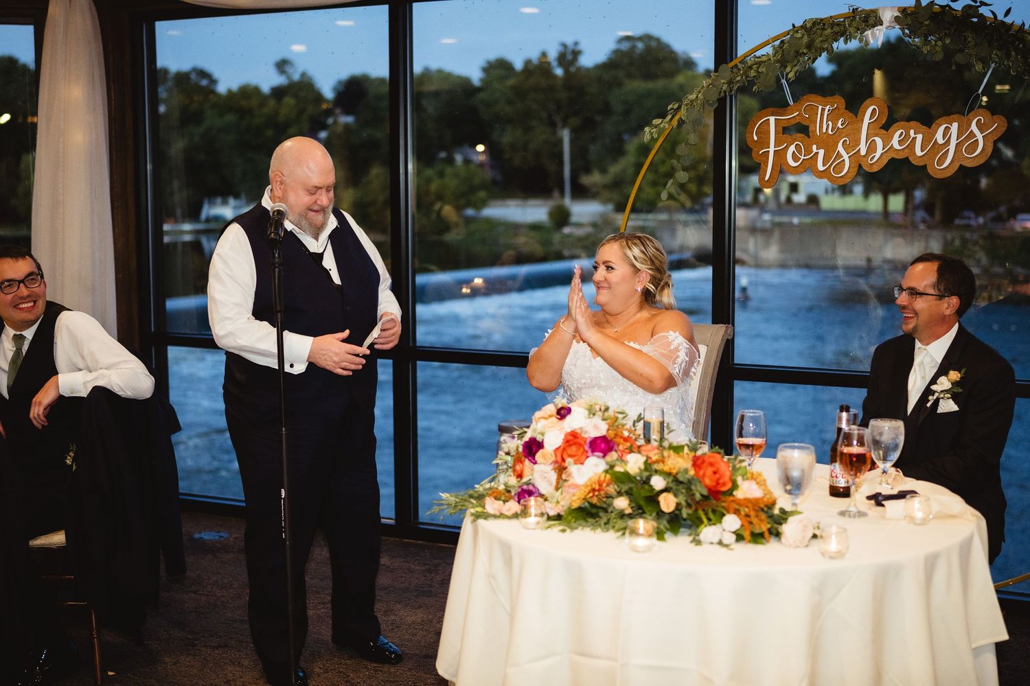 A wedding reception dining room along the waterfront at sunset with white tablecloths and floral centerpieces.