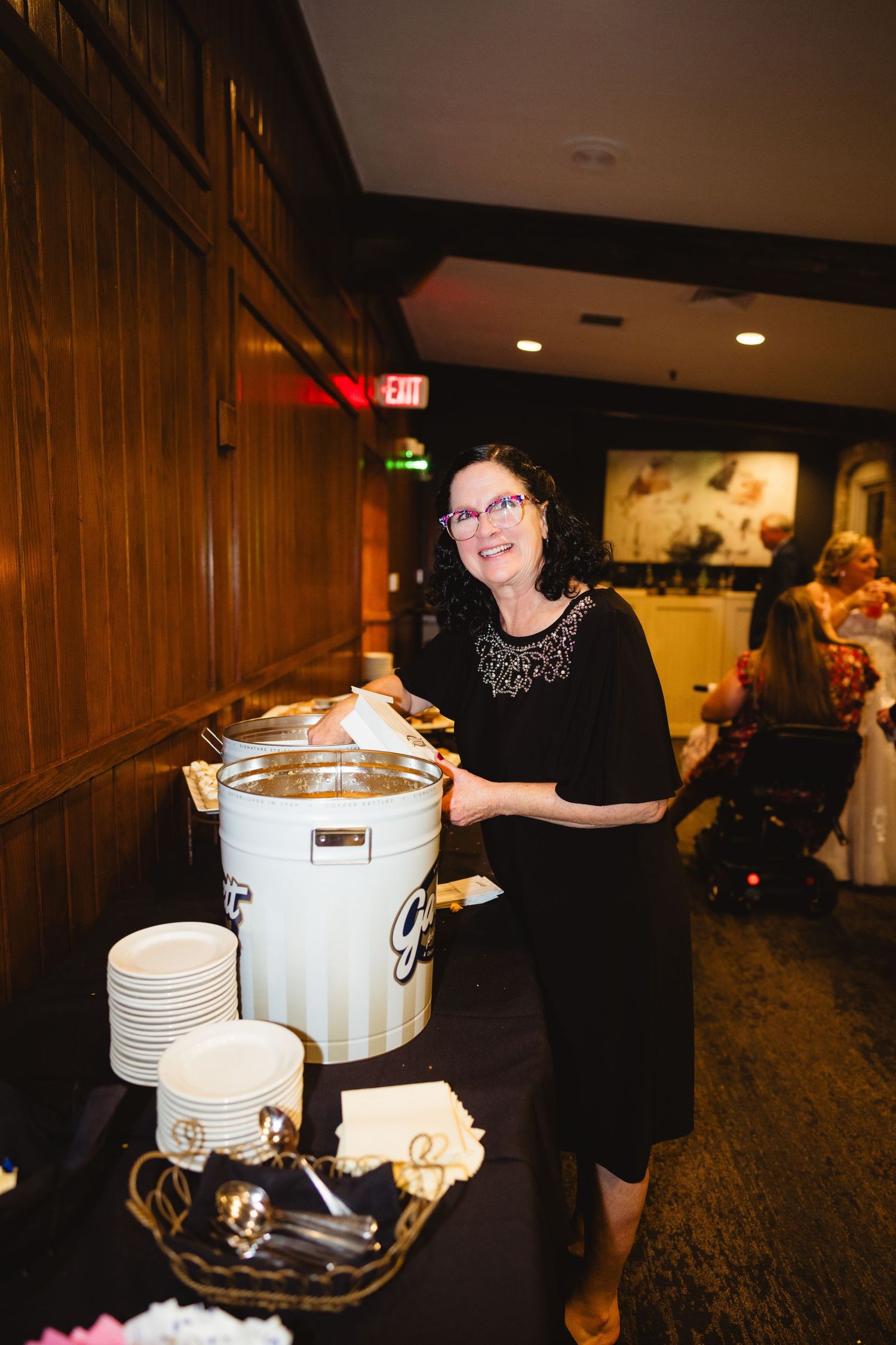 A woman in a black dress serves from a large white beverage dispenser at an indoor event.