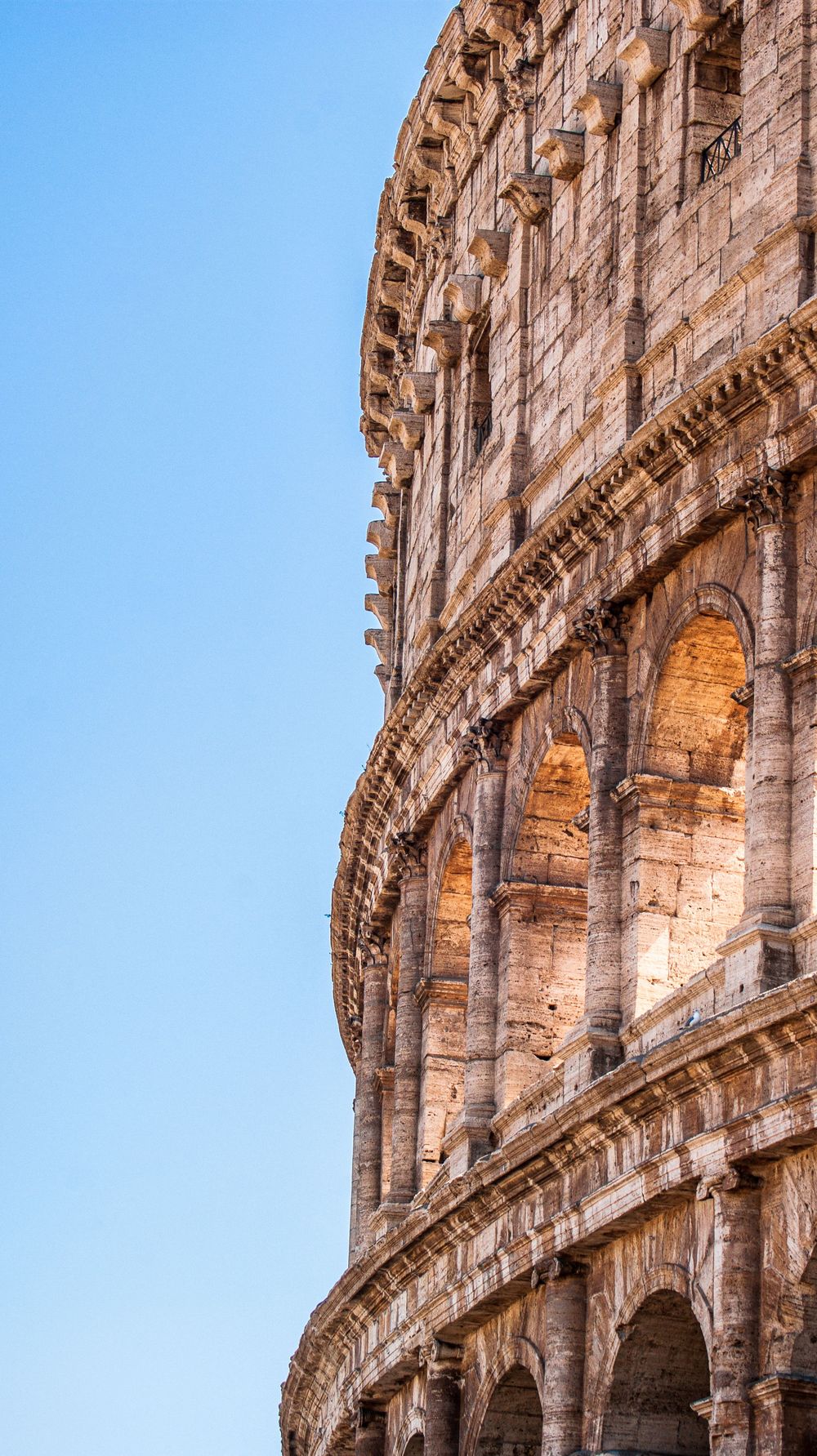 Ancient stone arches and architectural details of the Roman Colosseum against blue sky.