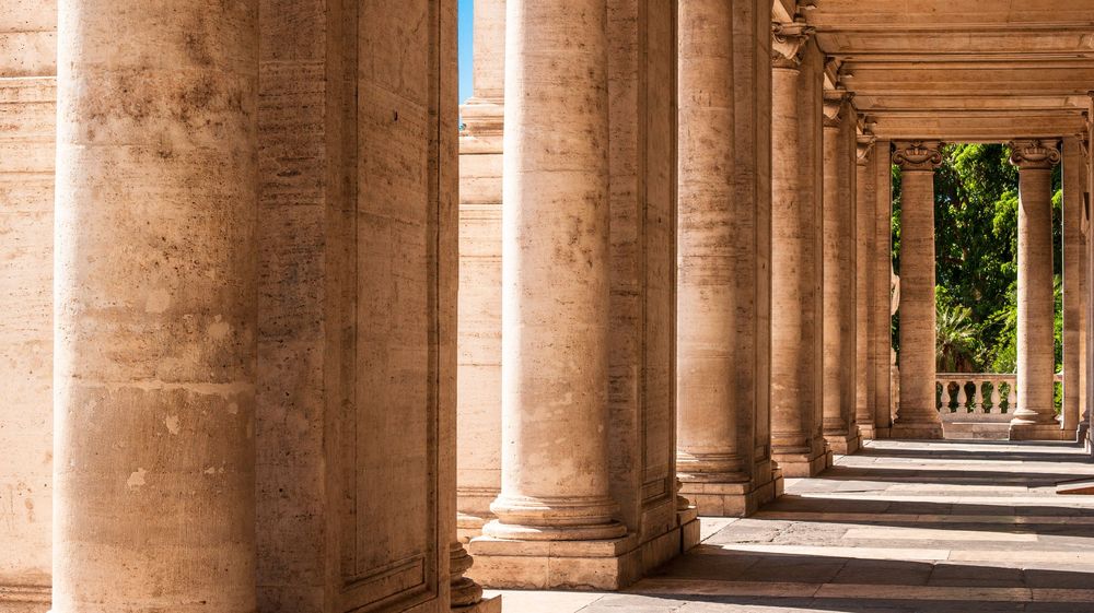 Historic stone colonnade with sunlit arched pillars creating long corridor perspective.