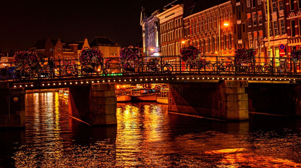 Nighttime cityscape reflecting on water with illuminated buildings and bridges creating golden reflections.