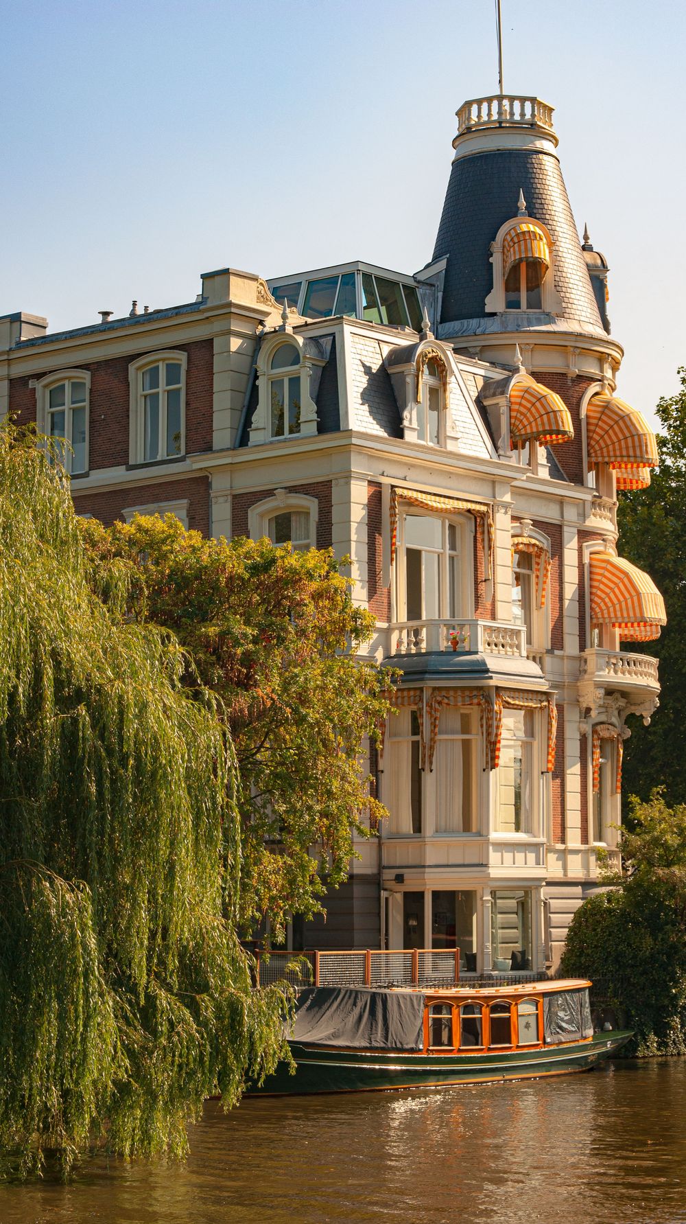 Elegant waterfront mansion with ornate towers and domes reflects in calm water with weeping willows along the shore.