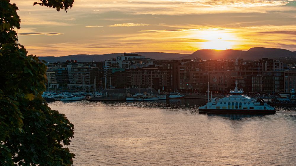 Panoramic sunset over harbor with boats and buildings silhouetted against orange sky.