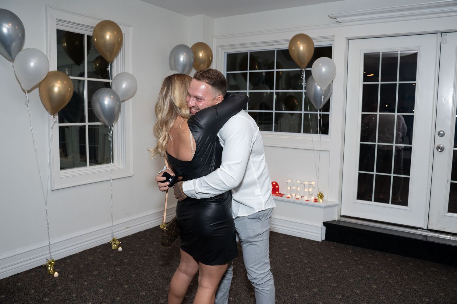 A couple embracing during a surprise proposal in a room with elegant balloon decorations.