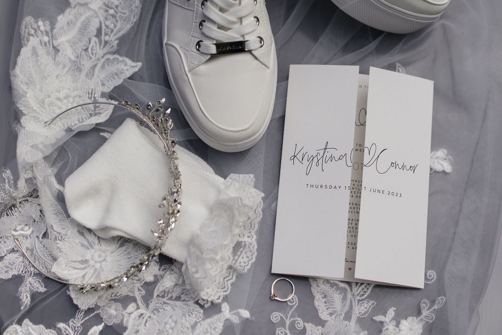 Wedding accessories including white shoes, lace garter, and invitations arranged on gray background.