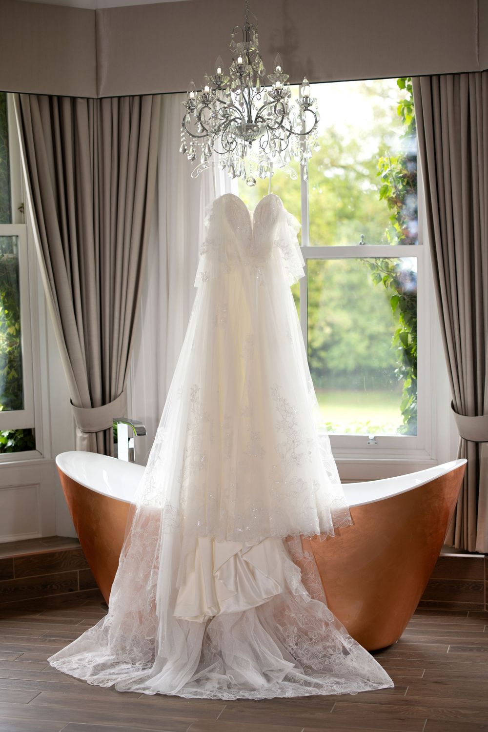 An elegant white wedding dress displayed in a room with gray curtains and natural lighting.