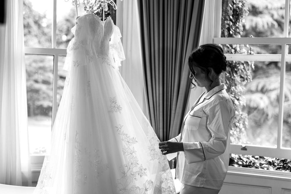 A beautiful wedding dress hanging in a bright room with floor-length curtains and natural sunlight.