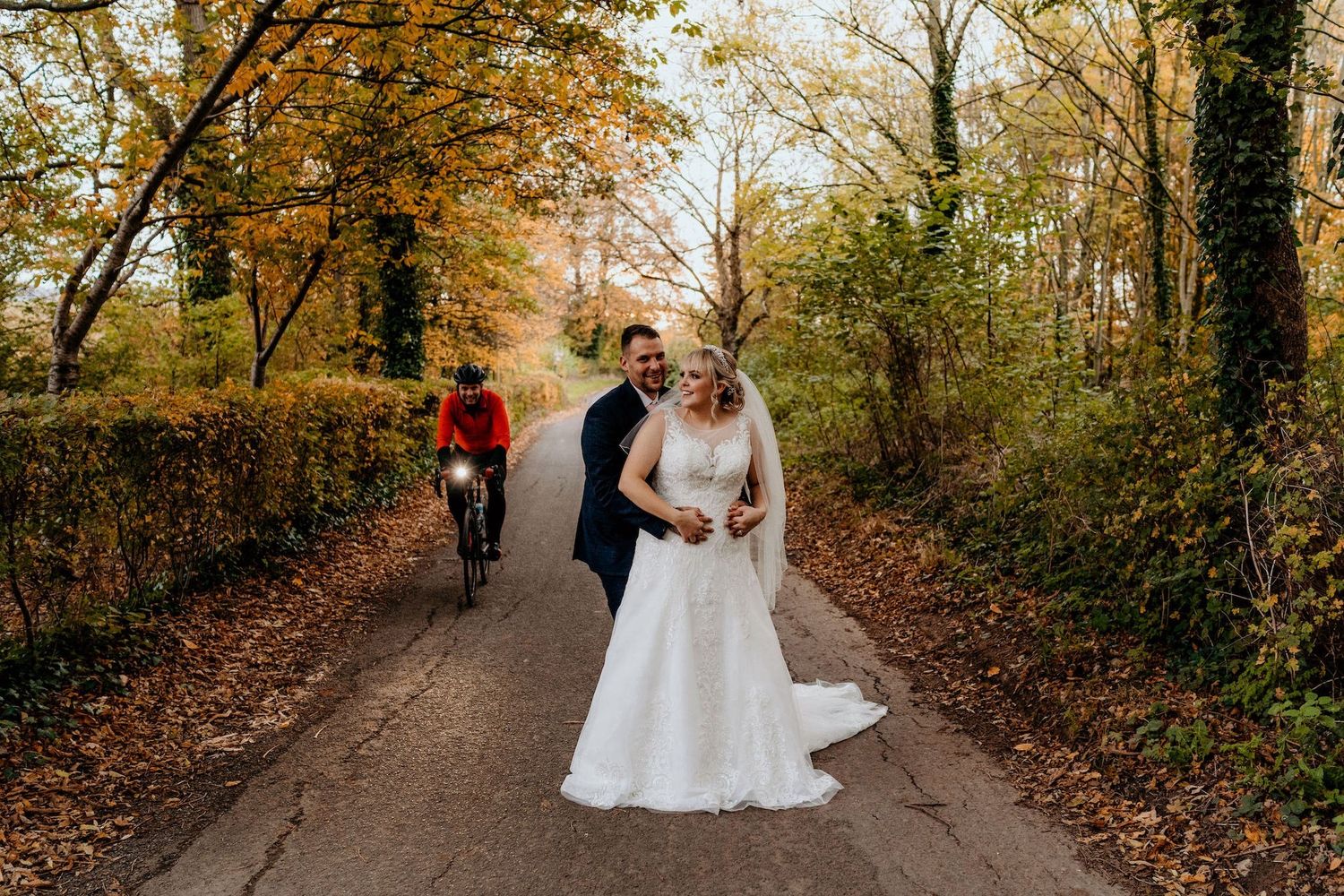 Couple in wedding attire walks along a scenic autumn path lined with golden-leaved trees and fallen foliage.