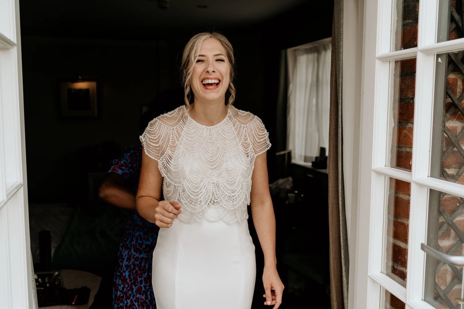 A bride wearing a lace cap sleeve wedding dress laughs joyfully by french doors in natural light.