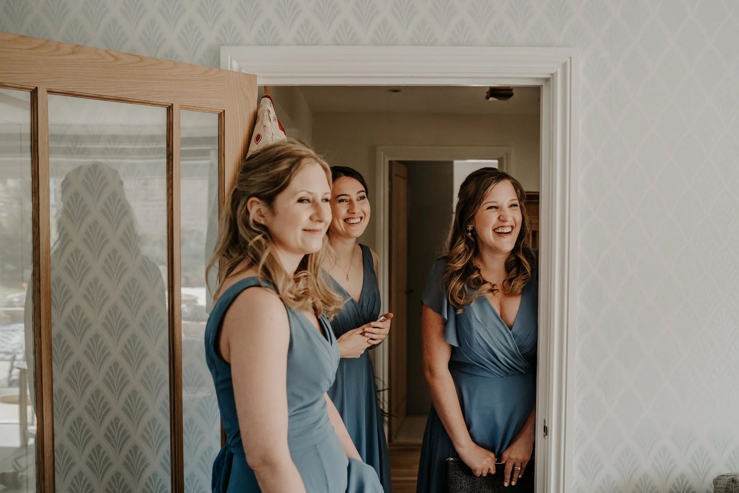 Group in blue gowns gather in room with geometric wallpaper during wedding preparations.