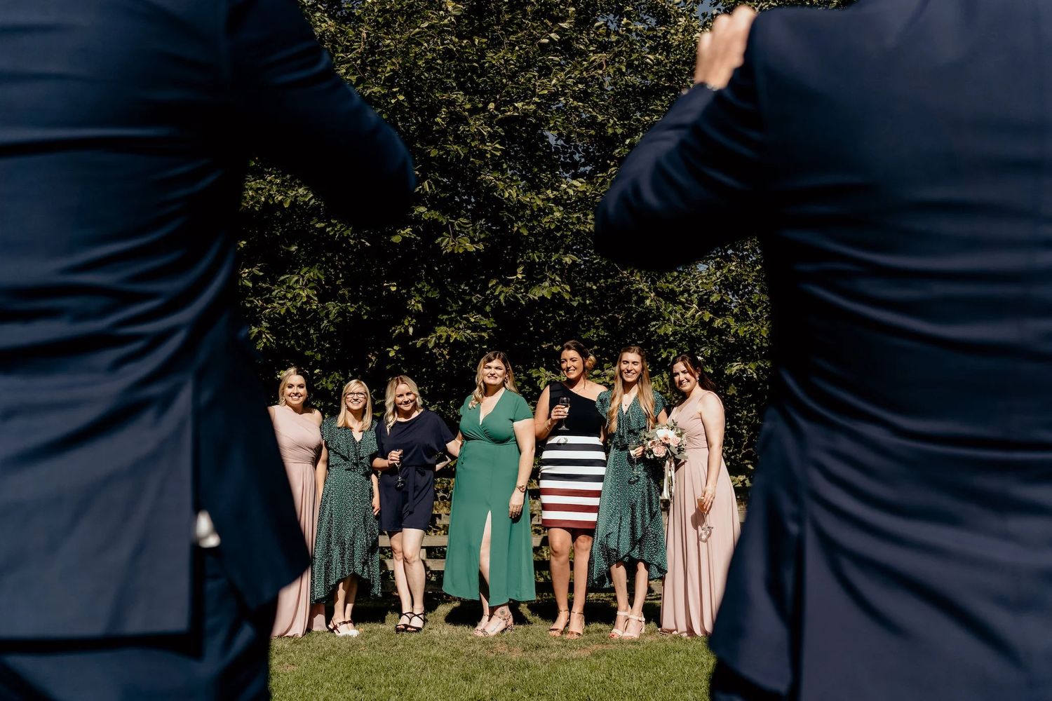 Wedding guests sit on grass beneath trees during an outdoor garden ceremony.