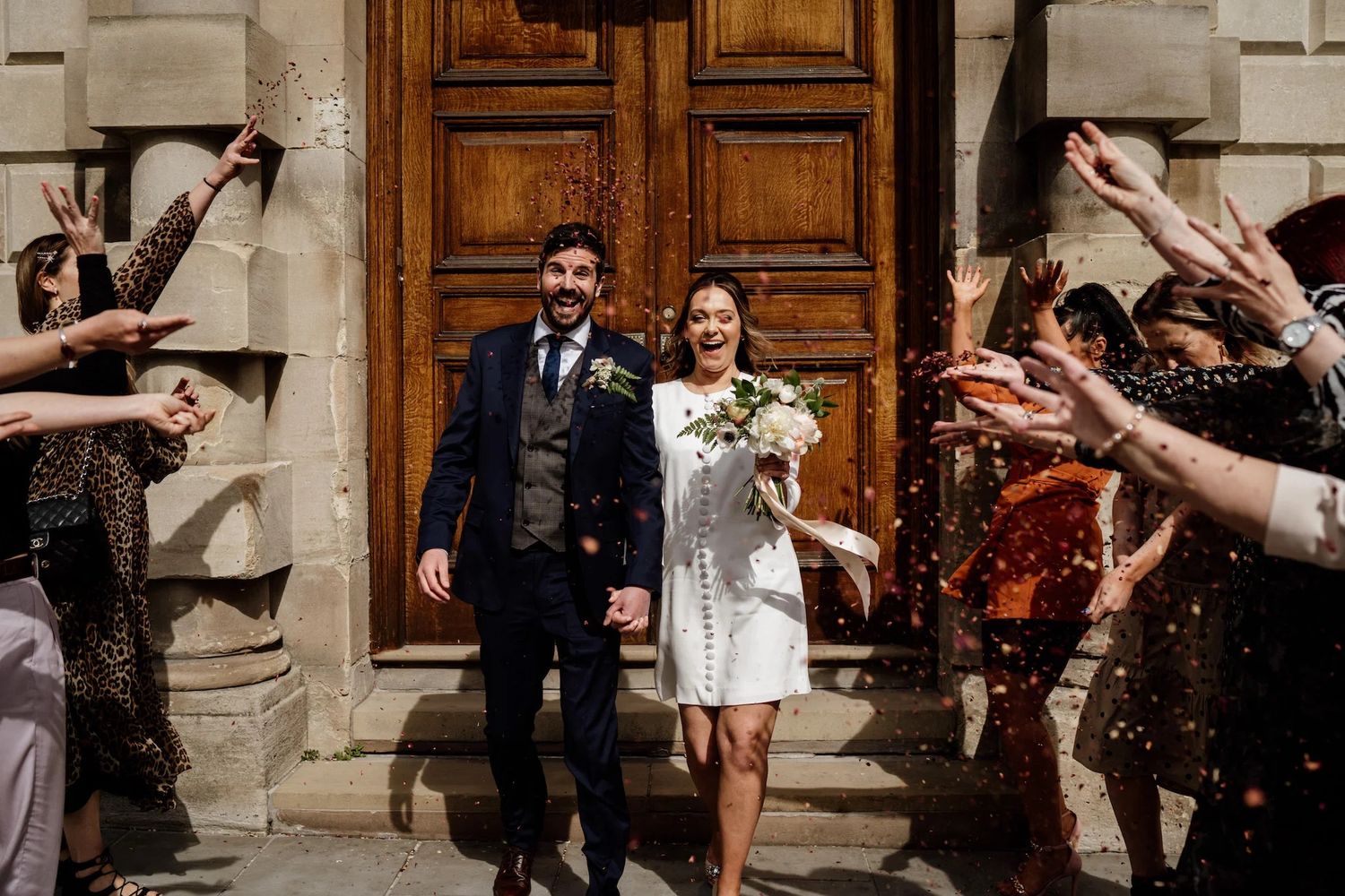 A couple exits a grand wooden doorway as guests shower them with confetti during a wedding celebration.