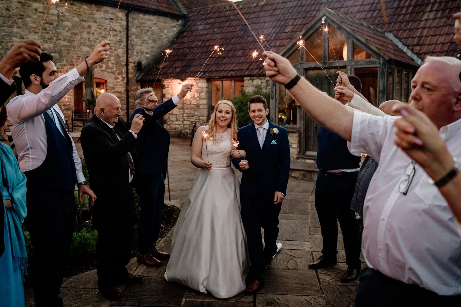 Wedding couple walks through sparkler tunnel held by guests outside rustic stone venue at dusk.