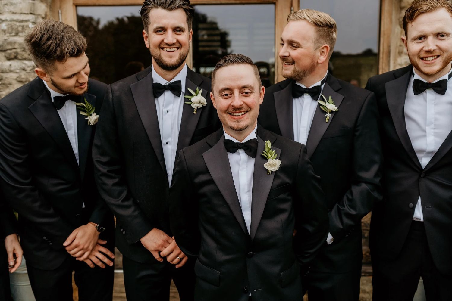 Groomsmen in black tuxedos and bowties smile together in a candid group photo.