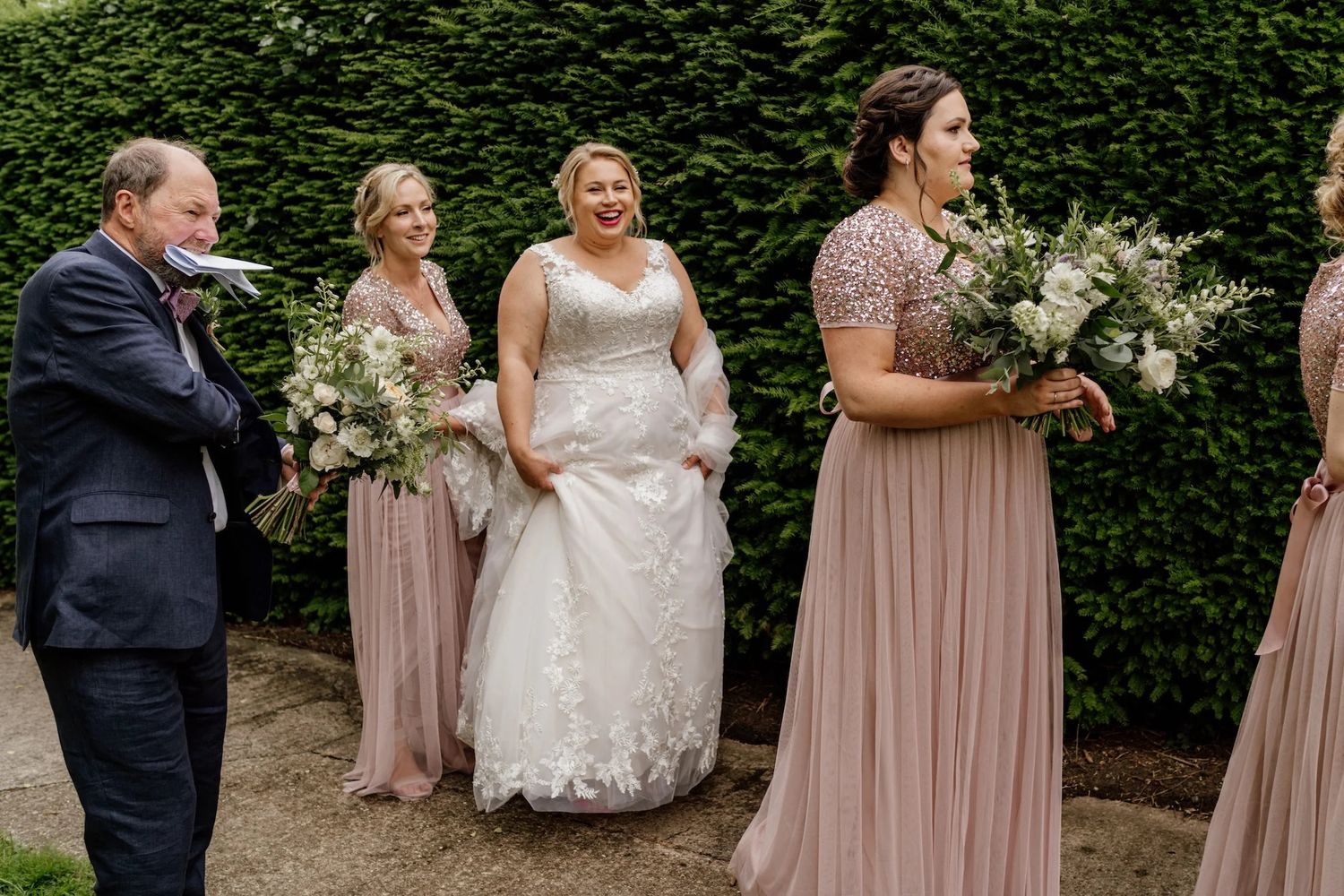 Bridal party in mauve dresses poses together surrounded by lush greenery at an outdoor wedding celebration.