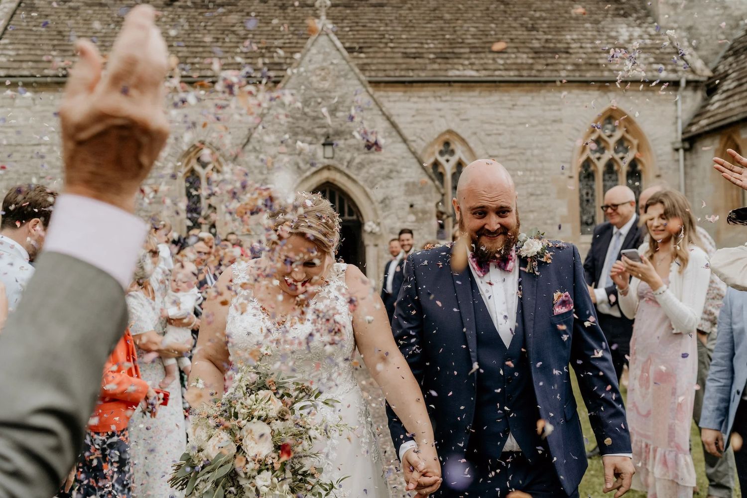 Wedding celebration with guests throwing confetti outside a rustic stone building.