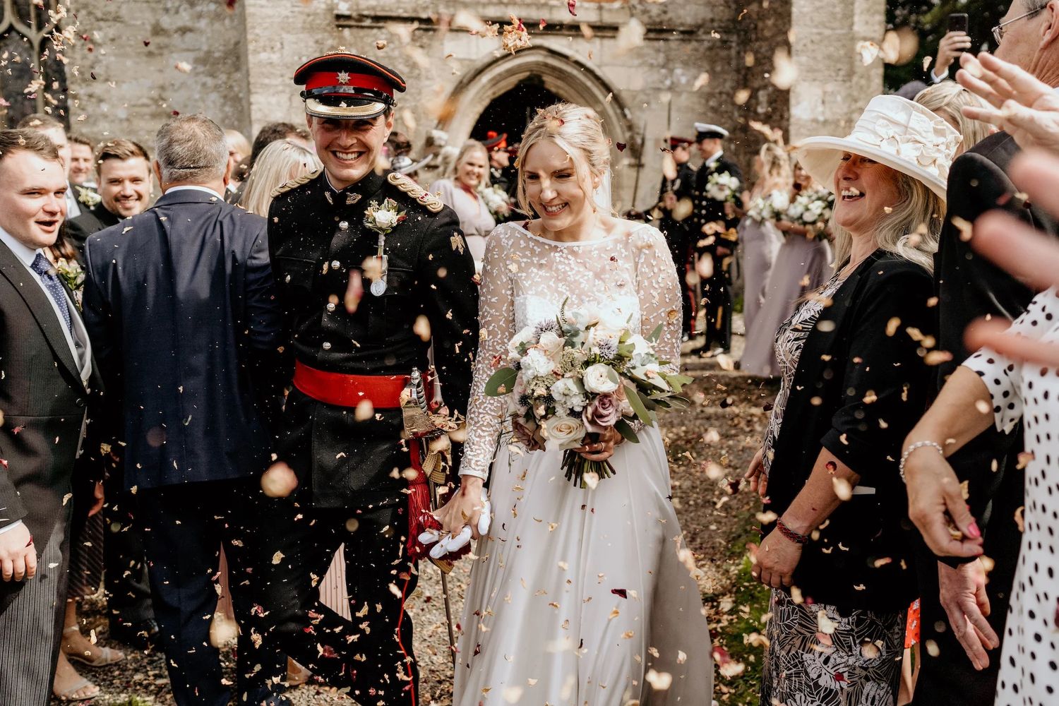 Wedding celebration with guests throwing confetti against a rustic stone wall backdrop.