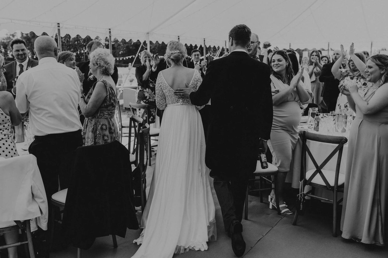 Black and white photo of wedding guests mingling during reception under draped tent.