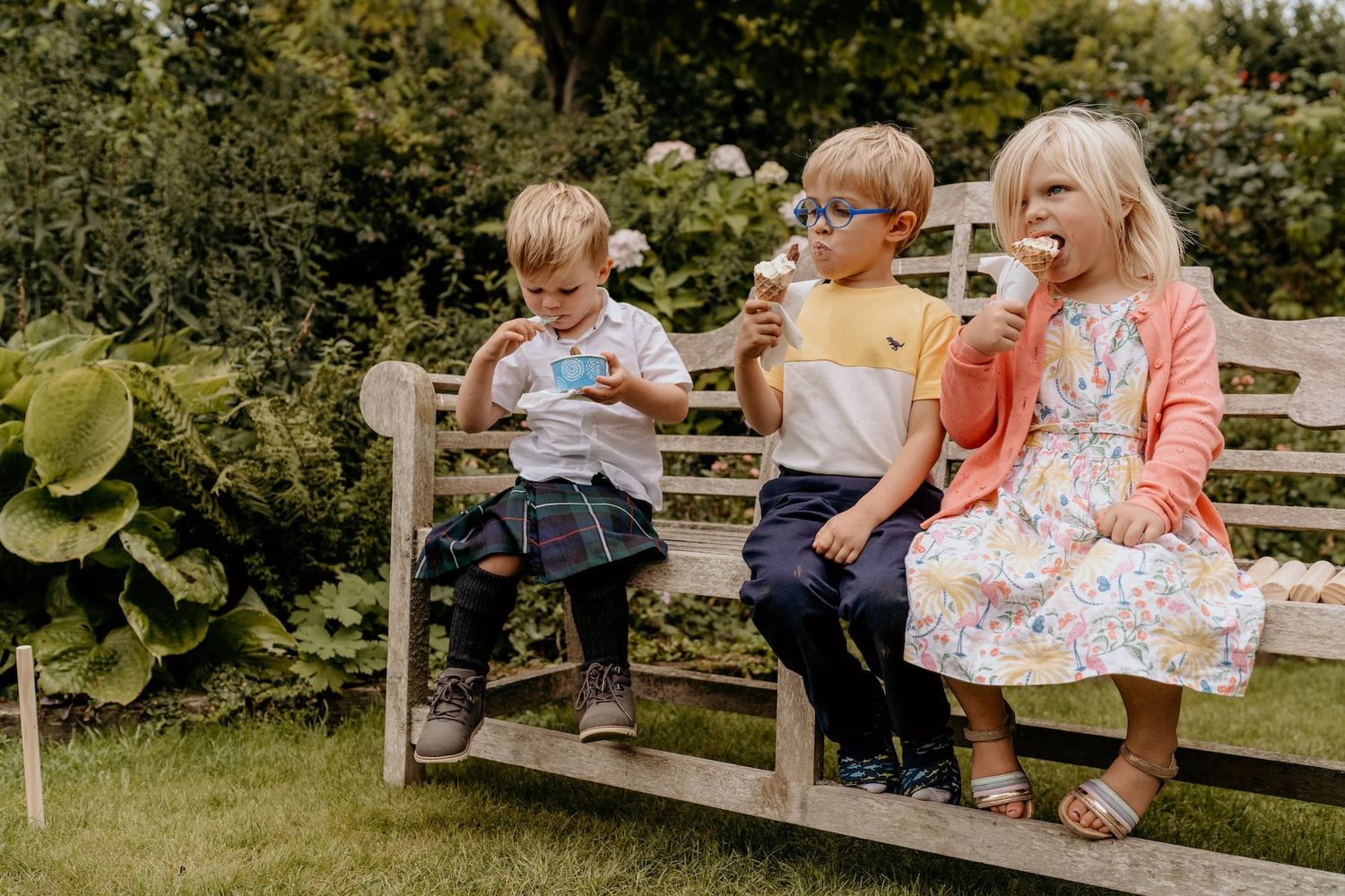 Children sit together on a wooden bench in a sunny garden setting during summer.