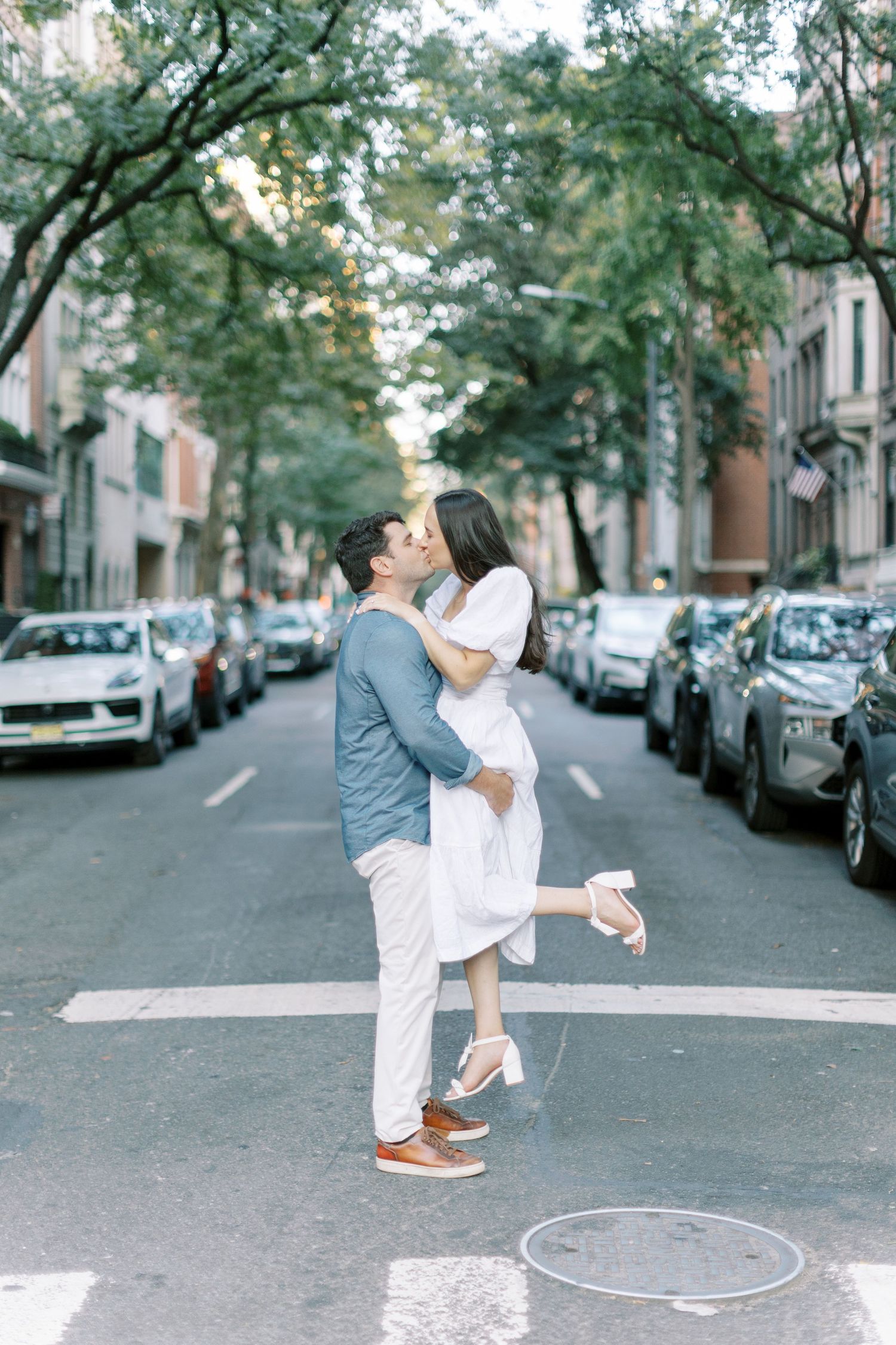 Two people dance playfully on a residential street lined with parked cars and trees in a romantic urban setting.