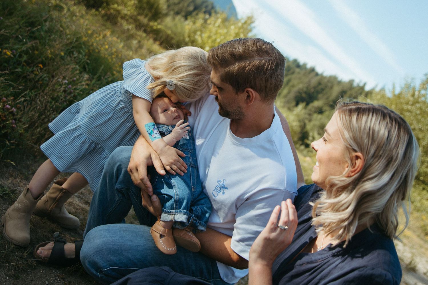 Family of three sitting outdoors, parents smiling at toddler in blue outfit, surrounded by lush greenery.
