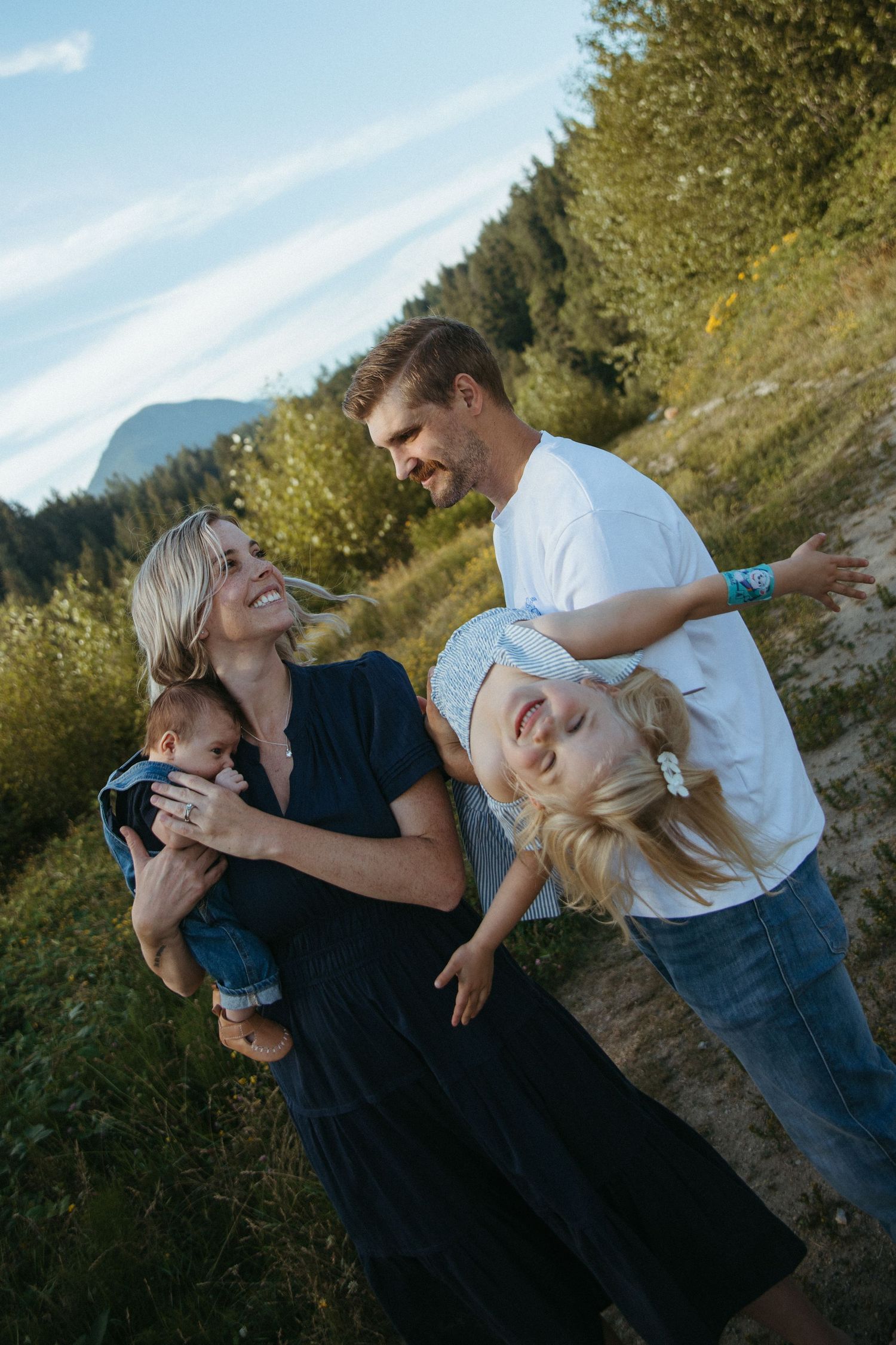 Happy family of four enjoying outdoor time, parents holding two young children in a scenic mountain meadow setting.