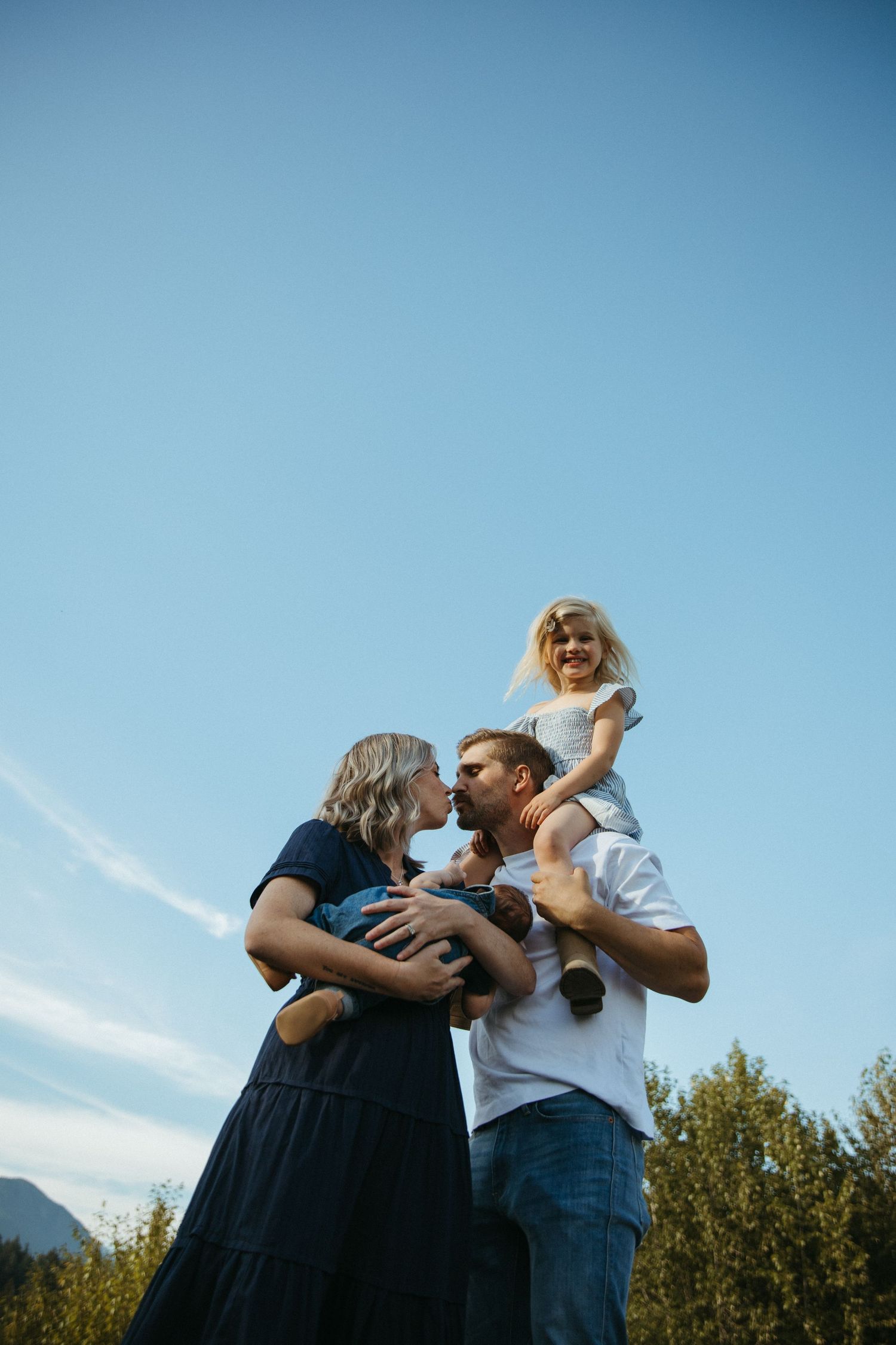 Happy family outdoors, parents kissing while dad carries daughter on shoulders against blue sky.