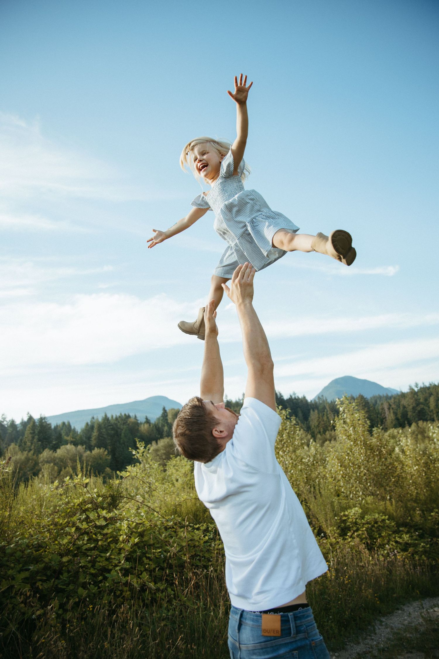 Father joyfully tosses laughing child into blue sky outdoors, mountains and green trees in background.