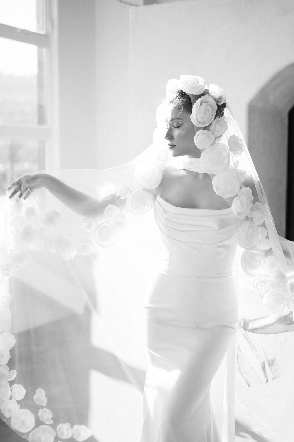 Elegant bride in fitted white gown and floral headpiece with veil stands gracefully in sunlit room.