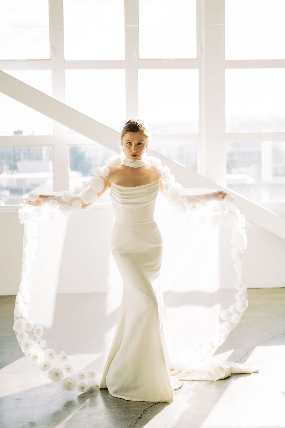 Bride in sleek white wedding gown stands in bright sunlit room with flowing veil dramatically spread wide.