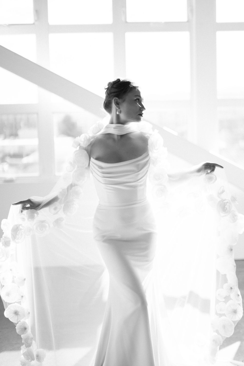 Artistic black and white silhouette of a bride in flowing dress against bright windows.